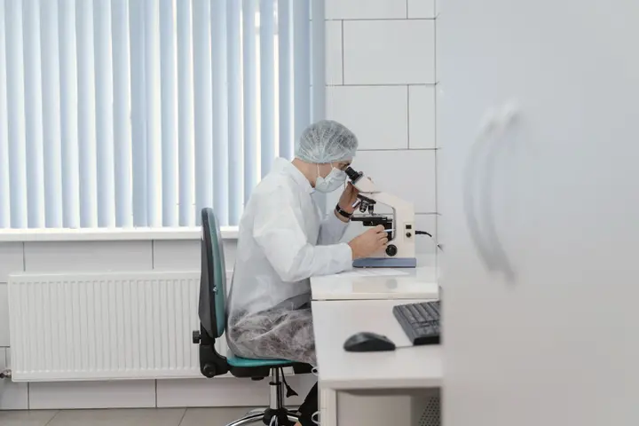 A clinician in a cap and gown works at a microscope in a clinic laboratory.