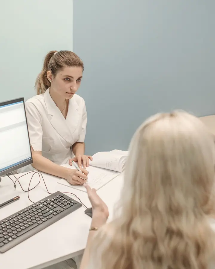 A doctor takes notes at her desk while a patient talks to her.