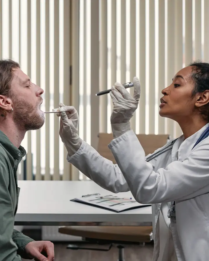 A doctor in a white coat checks a patient's throat in a bright clinic room.