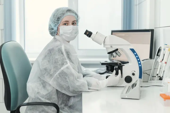 A clinician in a cap, mask and gown works at a microscope by a window.